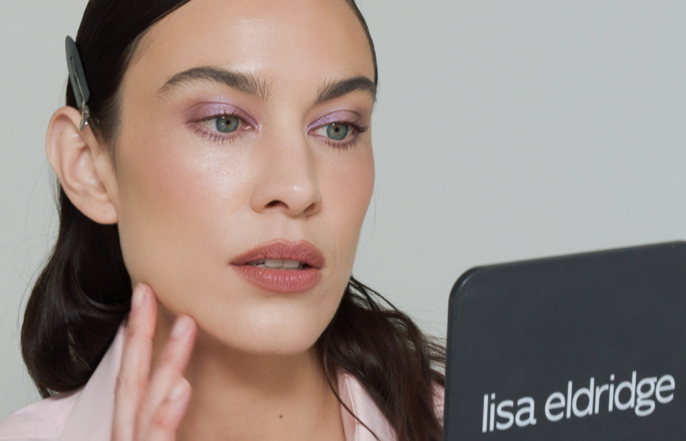 Alexa Chung holding a black mirror with 'Lisa Eldridge' branding, touching her jaw on a neutral background, wearing Betty Eyeshadow Palette shades.