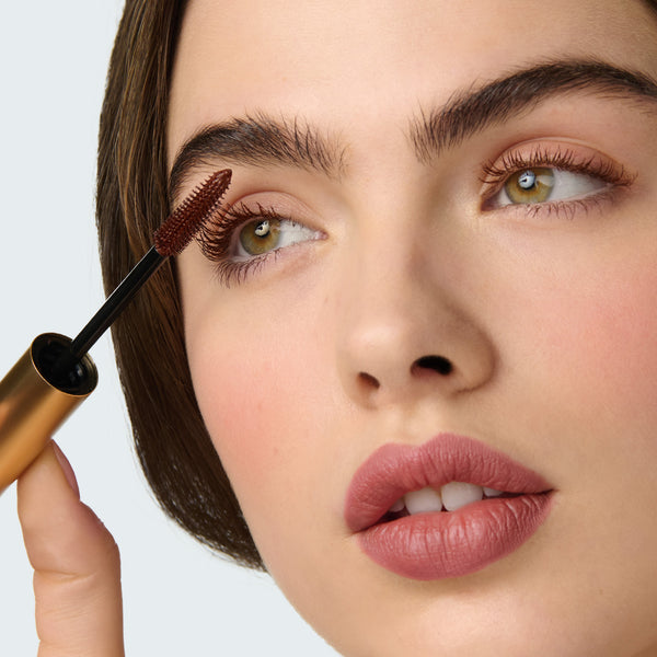Close-up of a model applying Kitten Lash Burnt Umber mascara to her eyelashes with a clear background