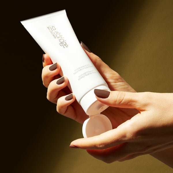 Hand squeezing the bottle of Seamless Skin Enhancing Treatment Cleanser  against a dark background