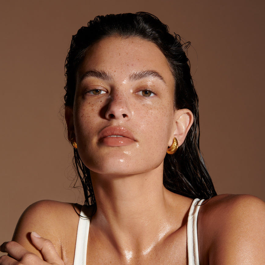 The head and shoulders of a model with glistening, tan skin. Their dark hair is wet and pushed back from their face and goes down their back. They have a natural makeup look on with a shine on their eyelids, nose, lips and chin. With Lisa Eldridge's Hero Ingredient Squalane