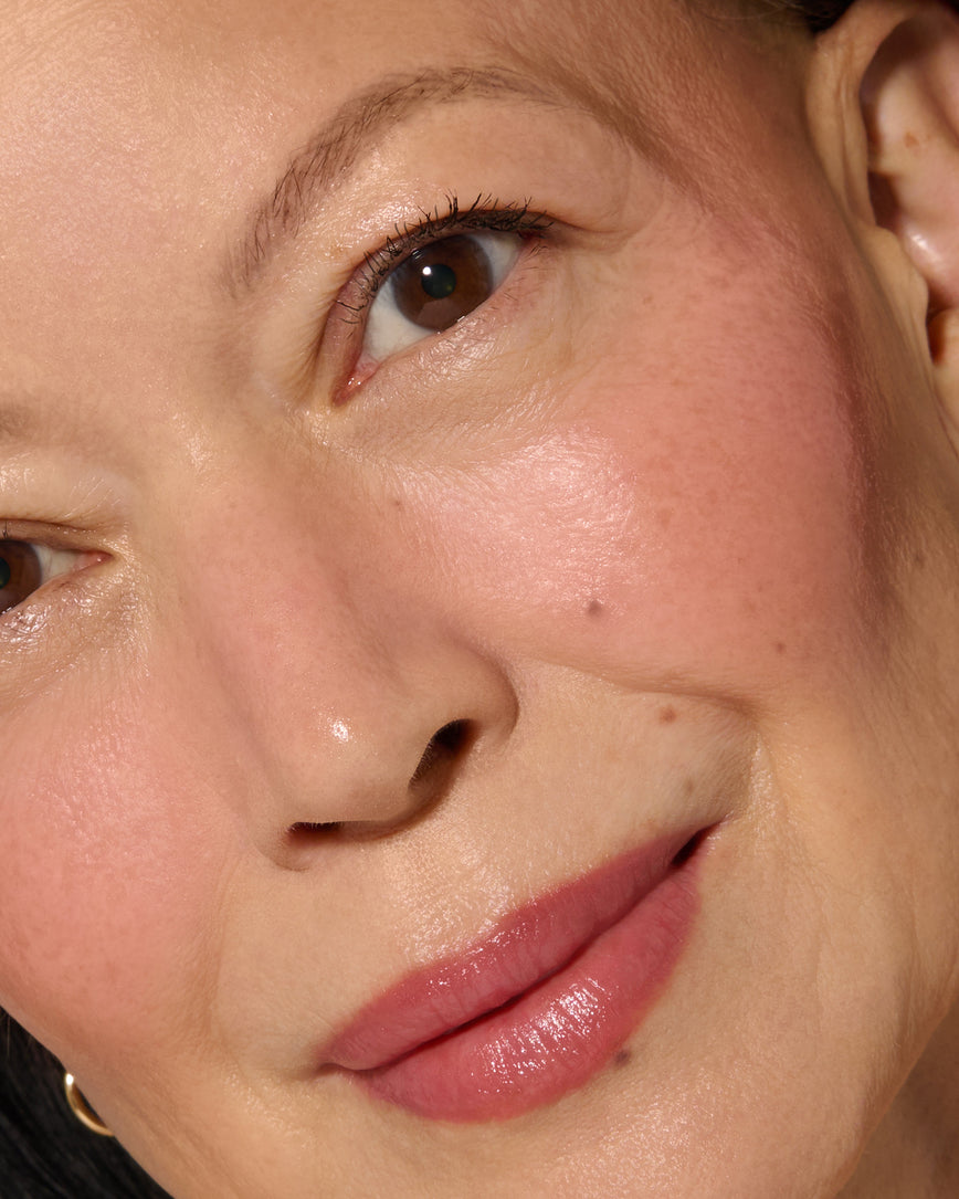 Close-up of a model's face with a neutral background, wearing Rose Madder Velatura Balm on lip and cheeks