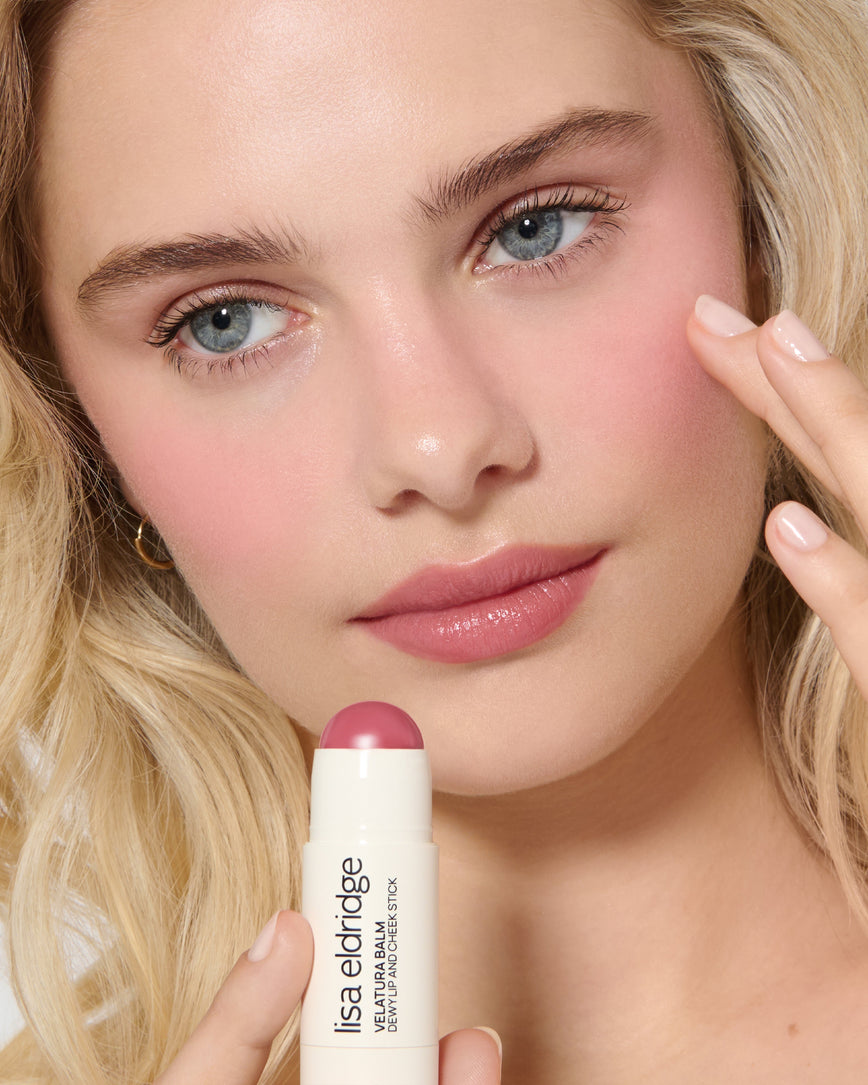 Close-up of a model's face with a neutral background, wearing Rose Madder Velatura Balm on lip and cheeks and applying the product to her lips