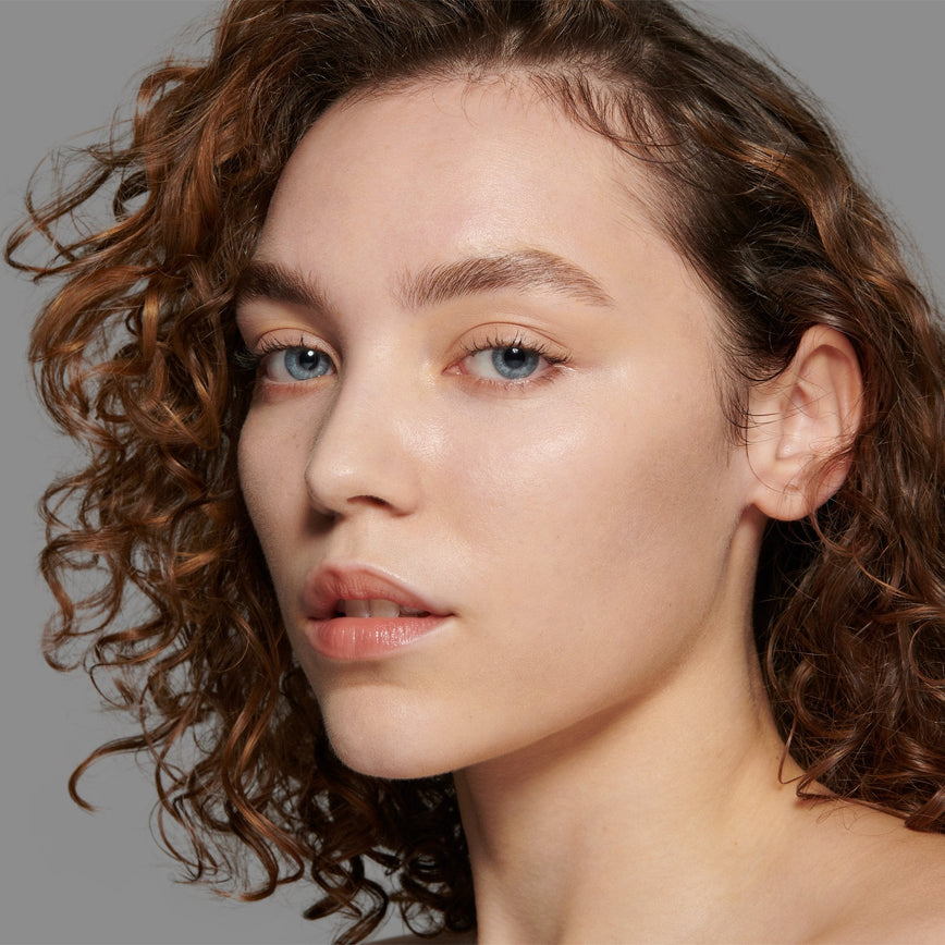 Lisa Eldridge Seamless Skin Foundation Pictured on Model. A young model with light skin and peach undertones, blue eyes and curly brown hair. The text says ‘No.4 With Peach Undertones'