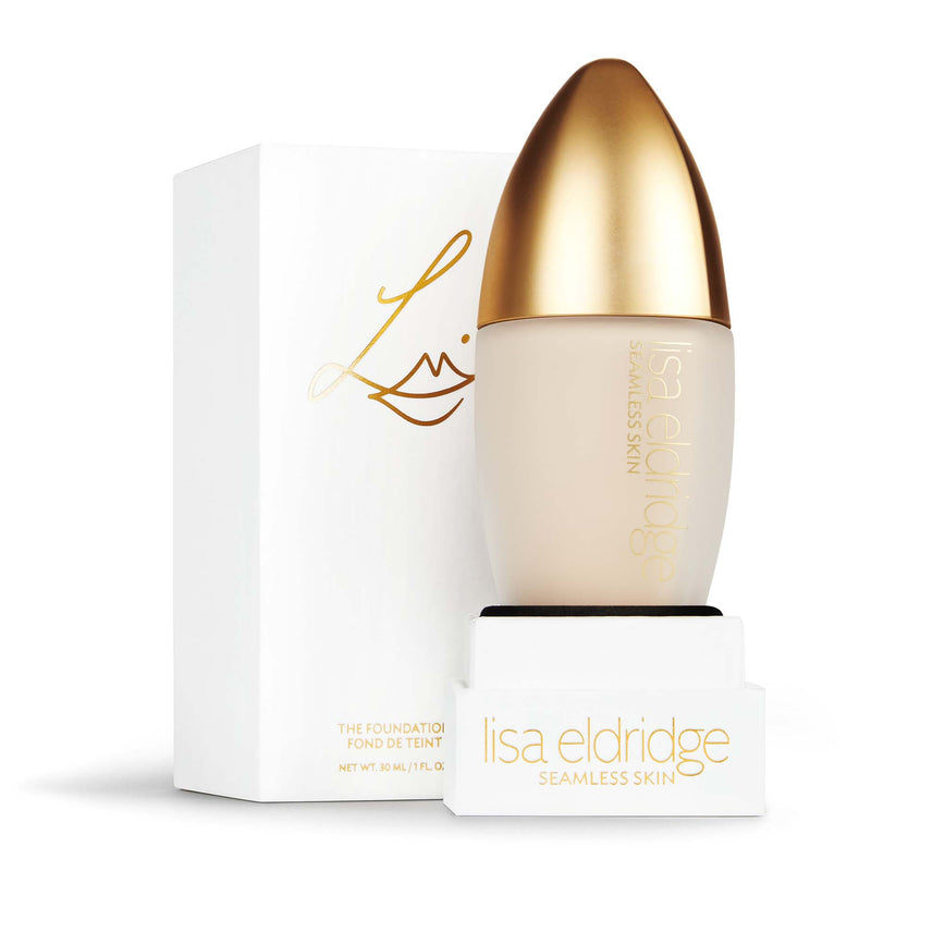 Lisa Eldridge Seamless Skin Foundation in a matte glass bottle with a gold lid. Behind the product is the box it comes packaged in. It is a white rectangular box with the Lisa eldridge logo in gold.