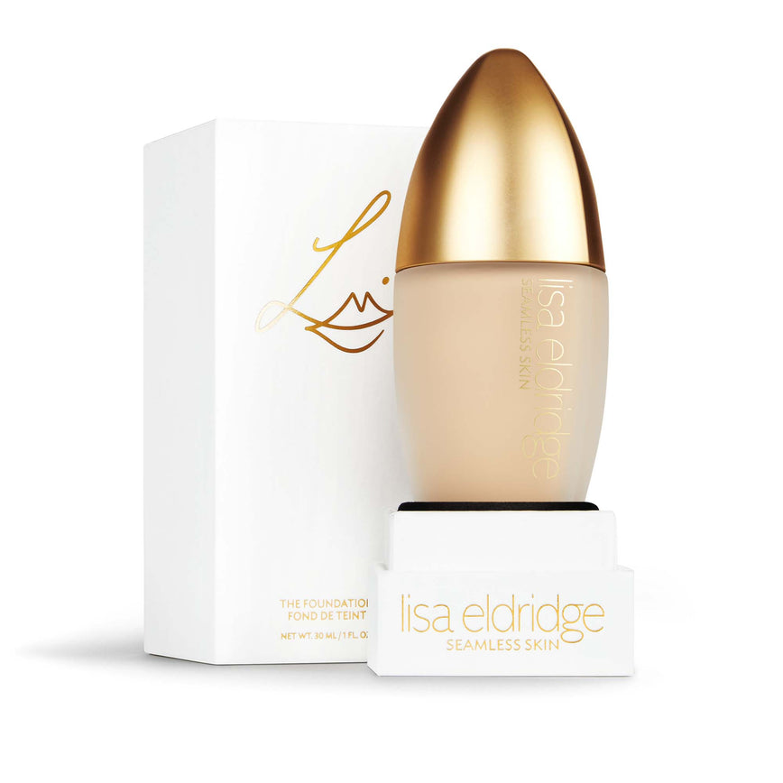 Lisa Eldridge Seamless Skin Foundation in a matte glass bottle with a gold lid. Behind the product is the box it comes packaged in. It is a white rectangular box with the Lisa Eldridge logo in gold.