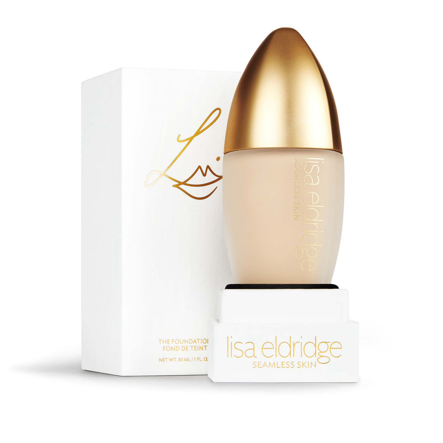 Lisa Eldridge Seamless Skin Foundation in a matte glass bottle with a gold lid. Behind the product is the box it comes packaged in. It is a white rectangular box with the Lisa Eldridge logo in gold.