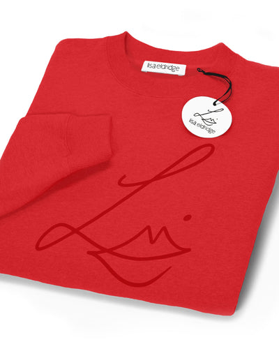 The Lisa Eldridge Studio Sweatshirt in red folded up. The crewneck and the cuffs have a fine rib. The Lisa Eldridge logo is large on the front. There is a label that says “lisa eldridge’ sewn on the inside. Attached to the size label is a circular card tag on a black ribbon with the Lisa Eldridge logo in gold