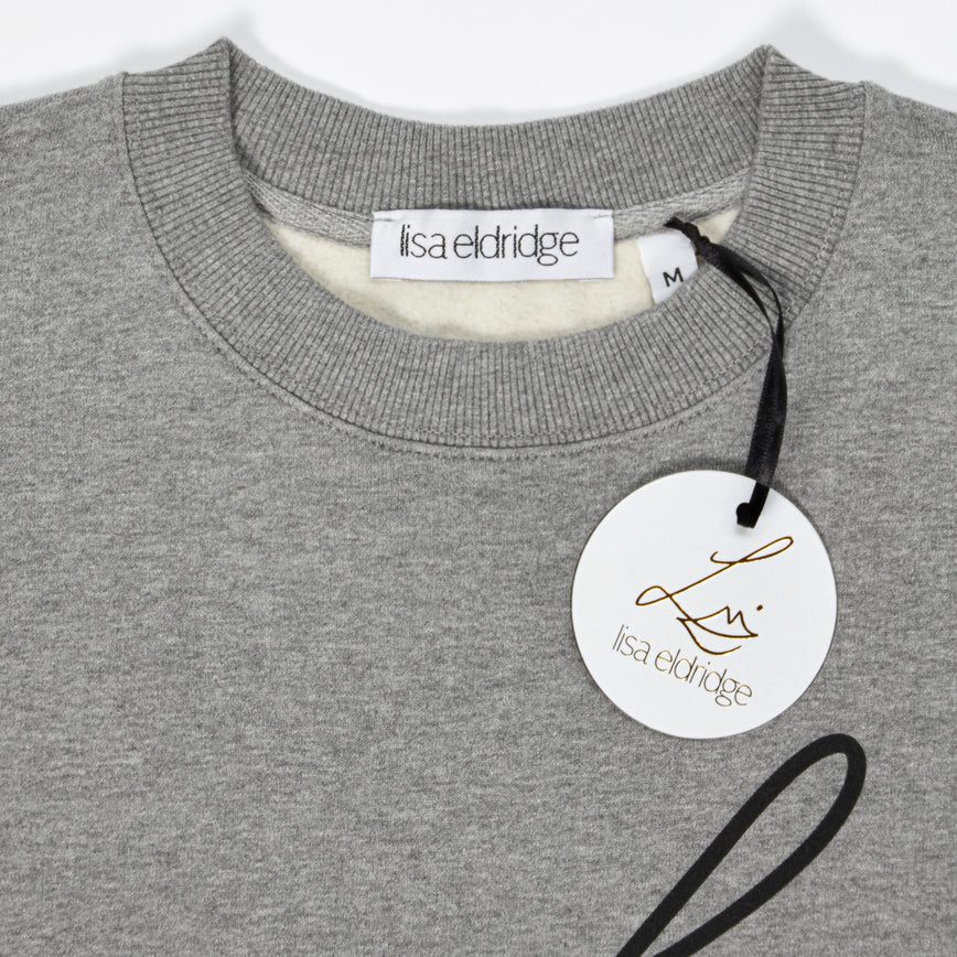 Close-up of the the Lisa Eldridge Studio Sweatshirt in grey. The crewneck has a fine rib. There is a label that says “lisa eldridge’ sewn on the inside. Attached to the size label is a circular card tag on a black ribbon with the Lisa Eldridge logo in gold.
